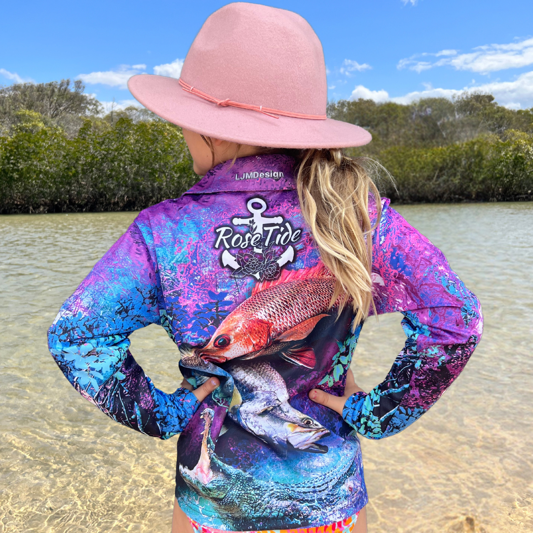 A girl wearing  A long sleeve fishing shirt with a vibrant, abstract background in purple, blue, and pink tones. The design features a barra, mangrove jack and a crab prominently on the front, along with the "Rose Tide" logo on the chest, which includes an anchor and rose