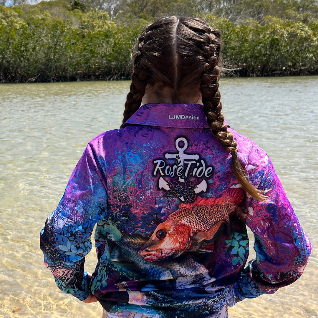 A girl wearing  A long sleeve fishing shirt with a vibrant, abstract background in purple, blue, and pink tones. The design features a barra, mangrove jack and a crab prominently on the front, along with the "Rose Tide" logo on the chest, which includes an anchor and rose