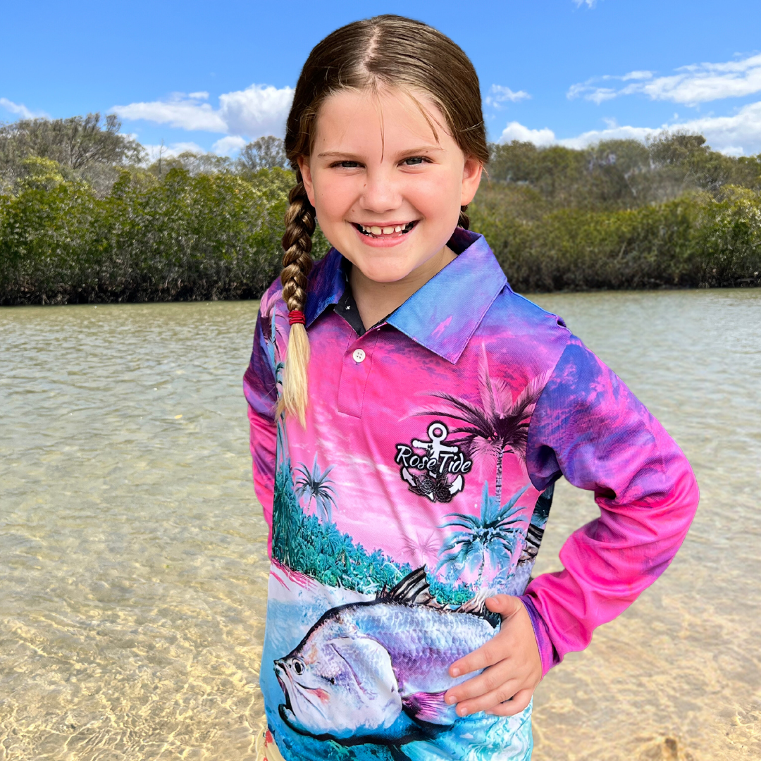 A girl wearing a long sleeve fishing shirt with a vibrant, tropical design featuring palm trees, a barra, and a crab in a colourful sunset beach scene. The shirt has the logo "Rose Tide" with an anchor and floral design on the back.