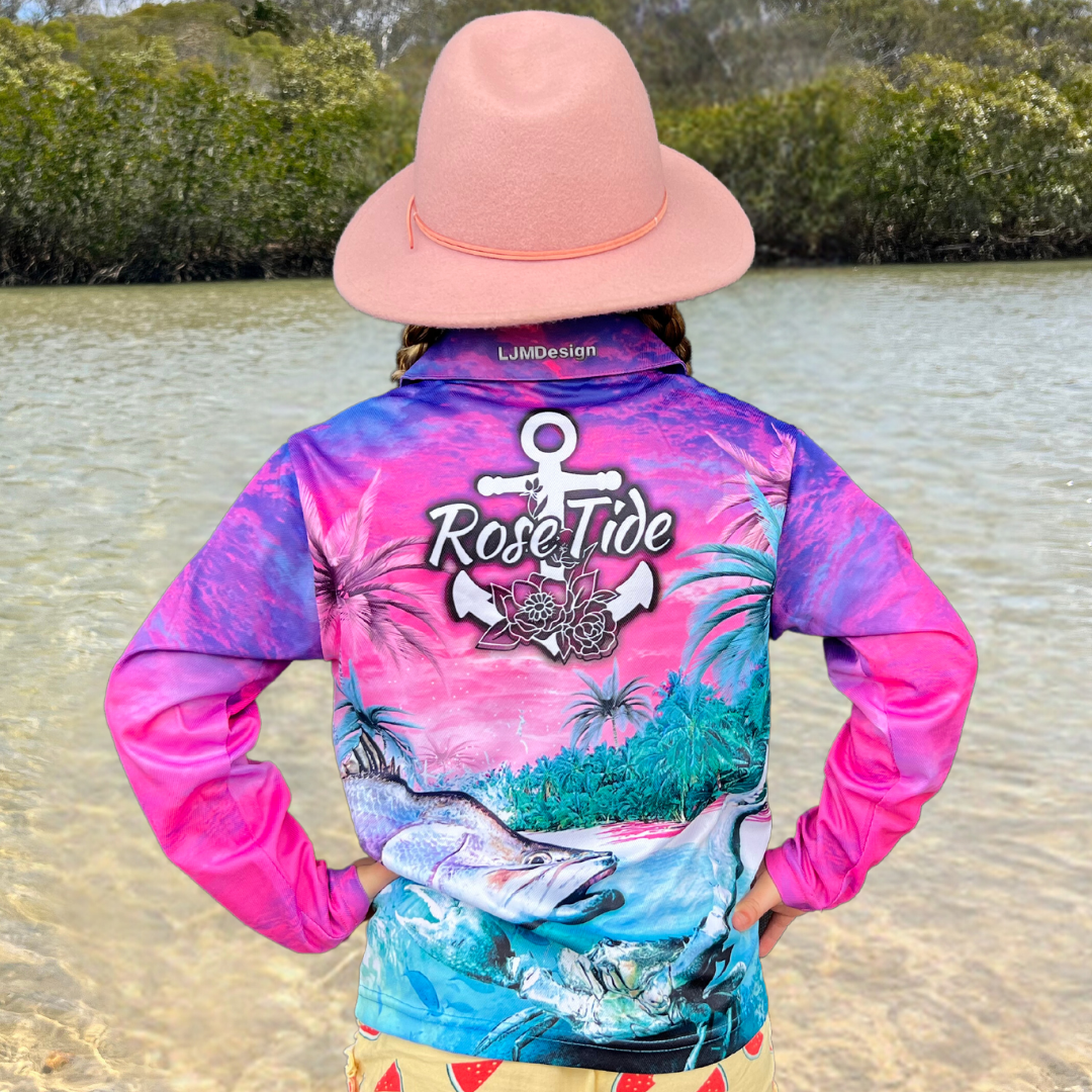 A girl wearing a long sleeve fishing shirt with a vibrant, tropical design featuring palm trees, a barra, and a crab in a colourful sunset beach scene. The shirt has the logo "Rose Tide" with an anchor and floral design on the back.