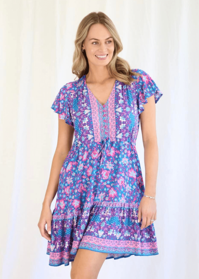 A Mini dress showcasing a vibrant blue and pink colour palette with intricate patterns. It has a playful, yet relaxed look that is likely versatile for various casual occasions. The v-neckline, flutter sleeves, and flounce at the bottom add a touch of femininity and ease to the overall design. The model wears the dress with a natural and confident stance, conveying a sense of comfort and style that the dress provides.