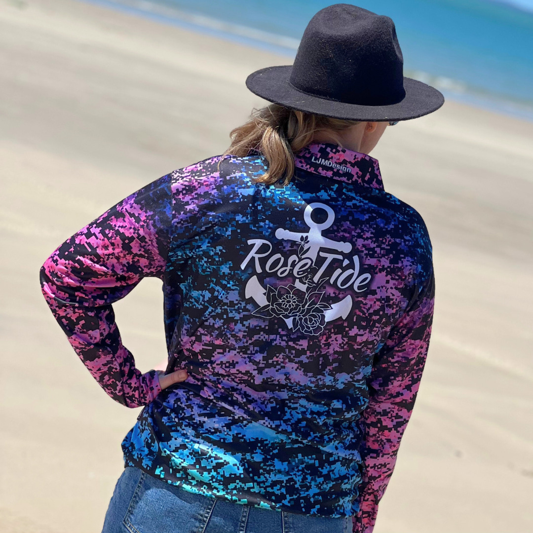 A long sleeve women's fishing shirt with a digital camouflage pattern in vibrant colors, including pink, purple, blue, and green. The shirt features the "Rose Tide" logo with an anchor and floral design on the chest. The overall design is bold and eye-catching.