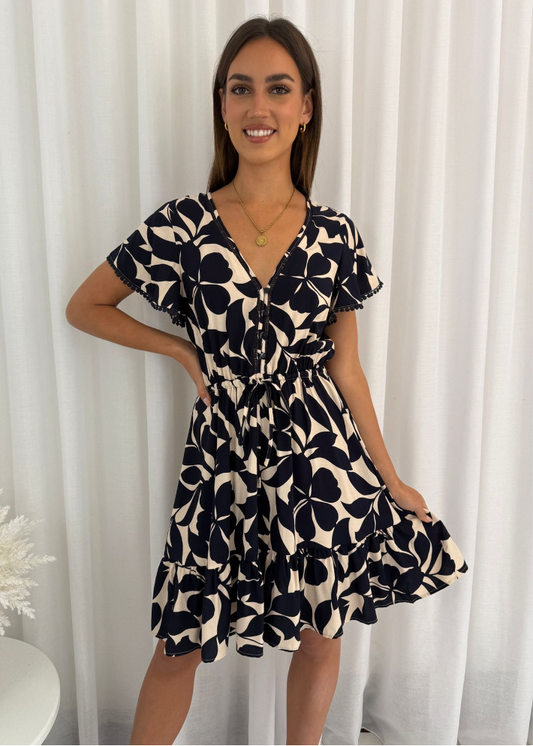 Model woman in the Celine Mini Dress, a summer boho-style navy blue and cream floral dress with a V-neckline, flutter sleeves, and a drawstring waist.