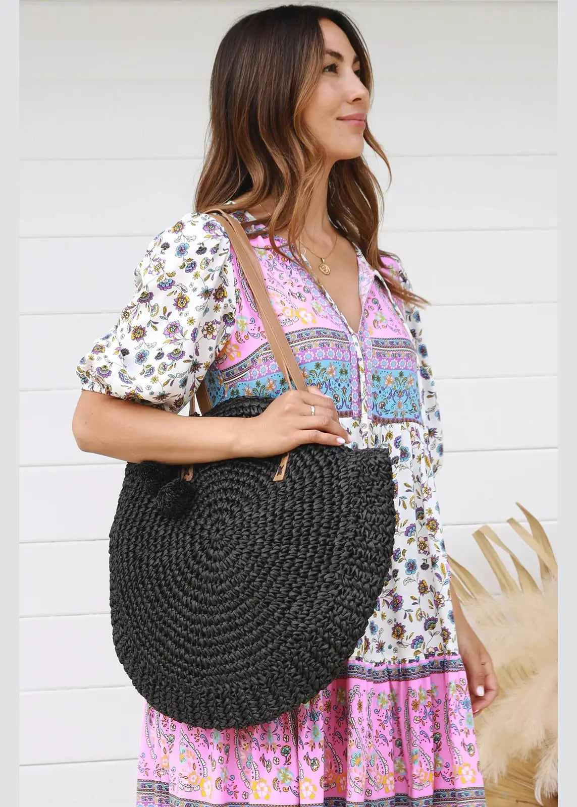 A woman holds a large, round, black, woven straw handbag with tan leather straps. The handbag features a circular, textured design and adds a chic, bohemian touch to her outfit. She is accessorized with a delicate gold necklace, and the background is a plain white wall, emphasizing the vibrant colours and textures of her attire and bag.