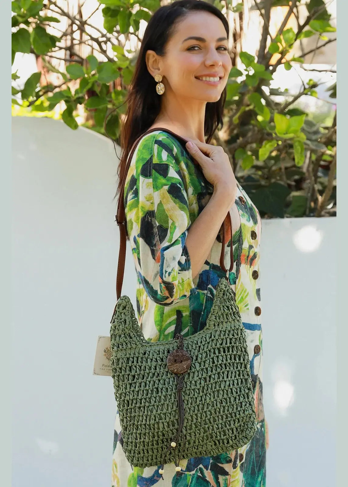 A woman is carrying a boho-style green, woven handbag with a wooden button closure and brown straps. 