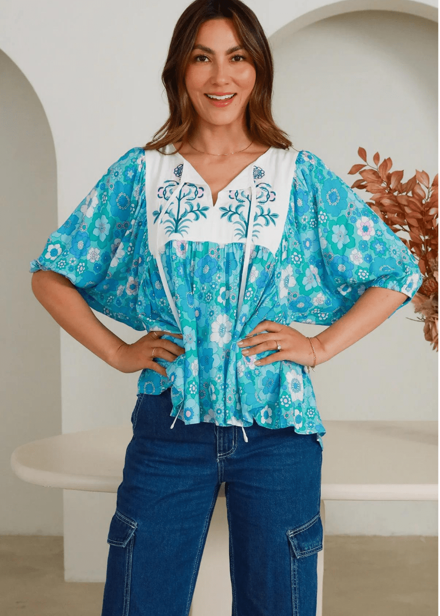 A blue and white floral embroidered top featuring a relaxed fit with flowing sleeves. The top has a V-neckline with intricate floral embroidery on the white, complemented by a vibrant blue floral print throughout the rest of the fabric. The model is wearing the top with blue jeans.