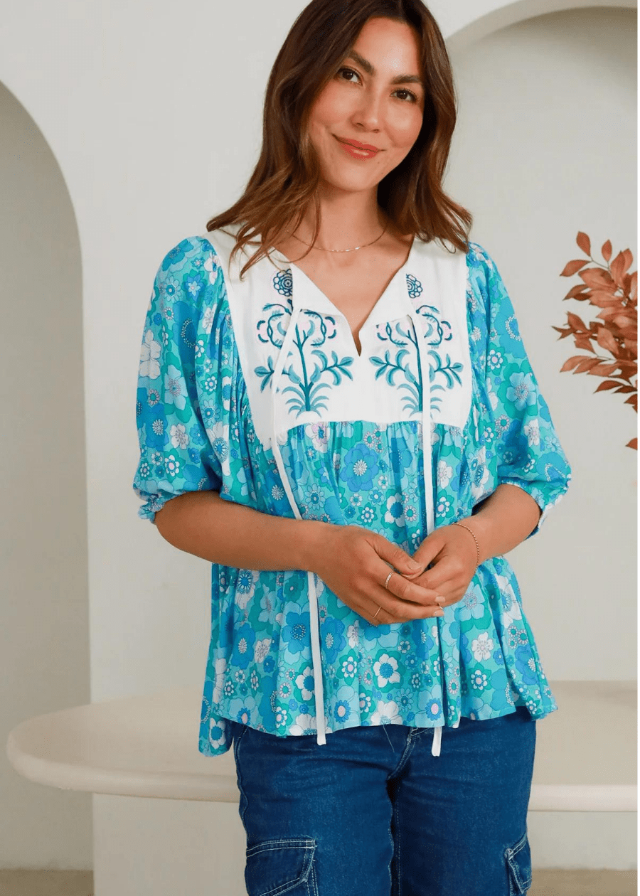 A blue and white floral embroidered top featuring a relaxed fit with flowing sleeves. The top has a V-neckline with intricate floral embroidery on the white yoke, complemented by a vibrant blue floral print throughout the rest of the fabric. The model is wearing the top with blue jeans.