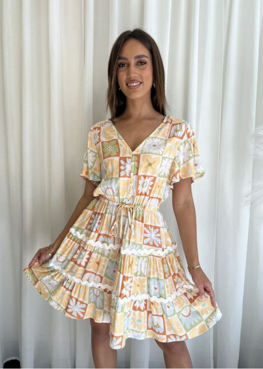 A smiling woman wearing a summer boho-style mini dress with soft yellow, orange, and pastel green floral and geometric patterns. The dress features a V-neckline, fluttery sleeves, an adjustable drawstring waist, and a tiered flared hem with delicate lace detailing, perfect for breezy summer outings.