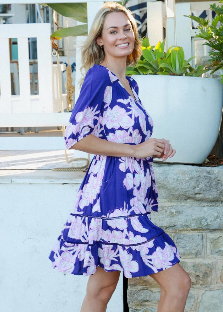 A woman wearing a blue mini dress with oversized pink floral print.  A flattering neckline that adds a touch of sophistication. Short sleeves with a puffed design for a stylish look. matching fabric belt to accentuate your waistline. Made from soft, breathable material, perfect for warm summer weather.