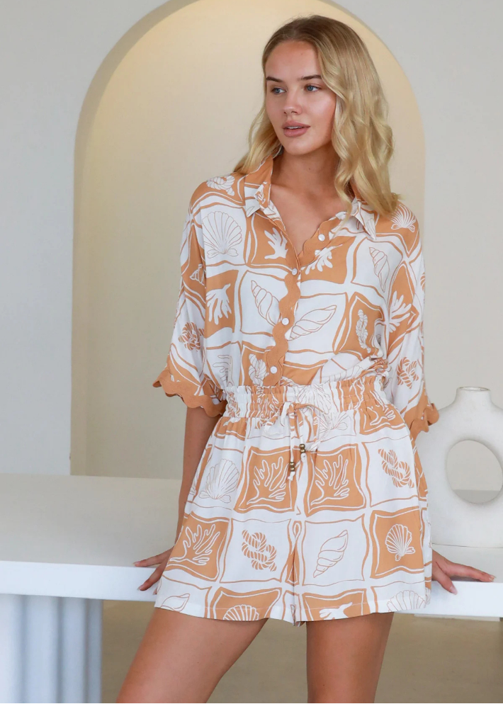 A woman is wearing the matching Summer Kai top and shorts, featuring nautical-inspired patterns and a casual, chic design. The coastal shell and coral print in beige and white showcases an elastic waistband and relaxed fit.