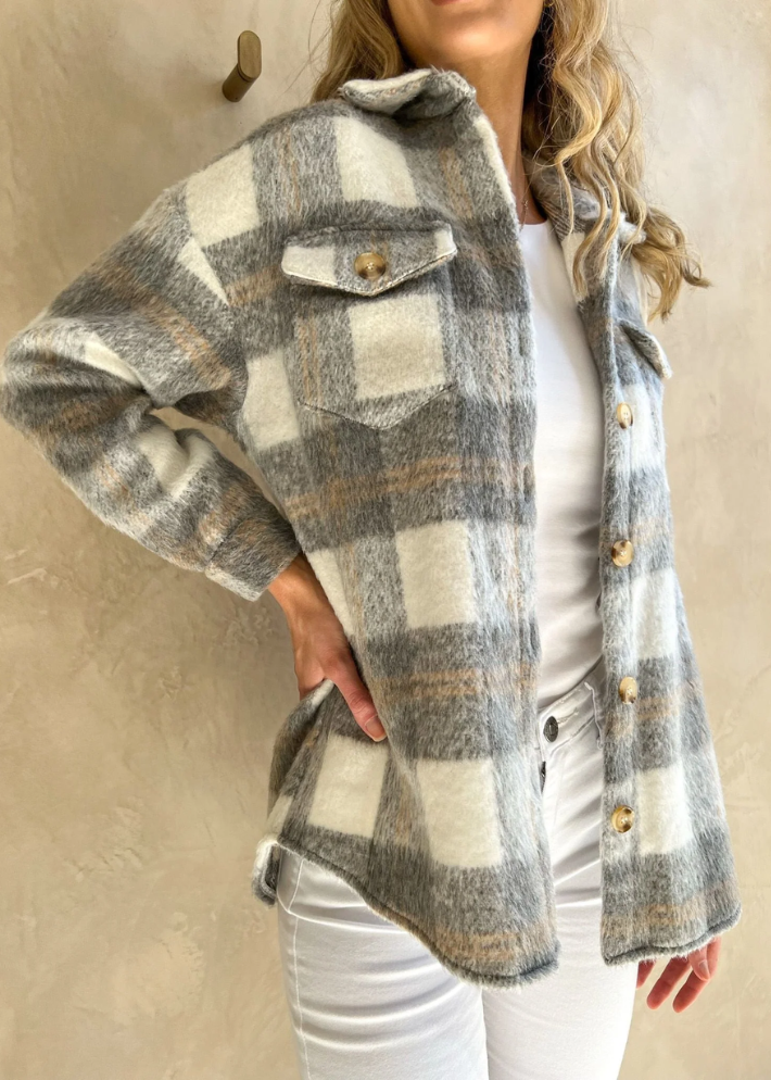 A woman wearing a relaxed-fit plaid jacket in shades of grey and white, featuring button details and front pockets. She paired the jacket with a white top and white high-waisted pants, creating a cozy and stylish outdoor look.