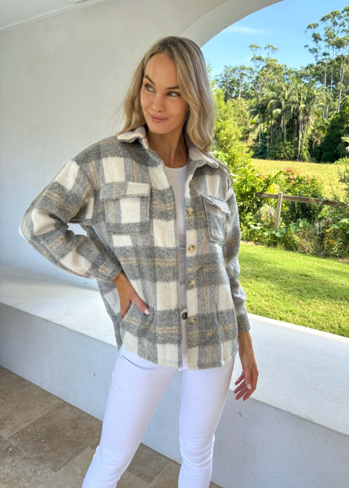 A woman wearing a relaxed-fit plaid jacket in shades of grey and white, featuring button details and front pockets. She paired the jacket with a white top and white high-waisted pants, creating a cozy and stylish outdoor look.