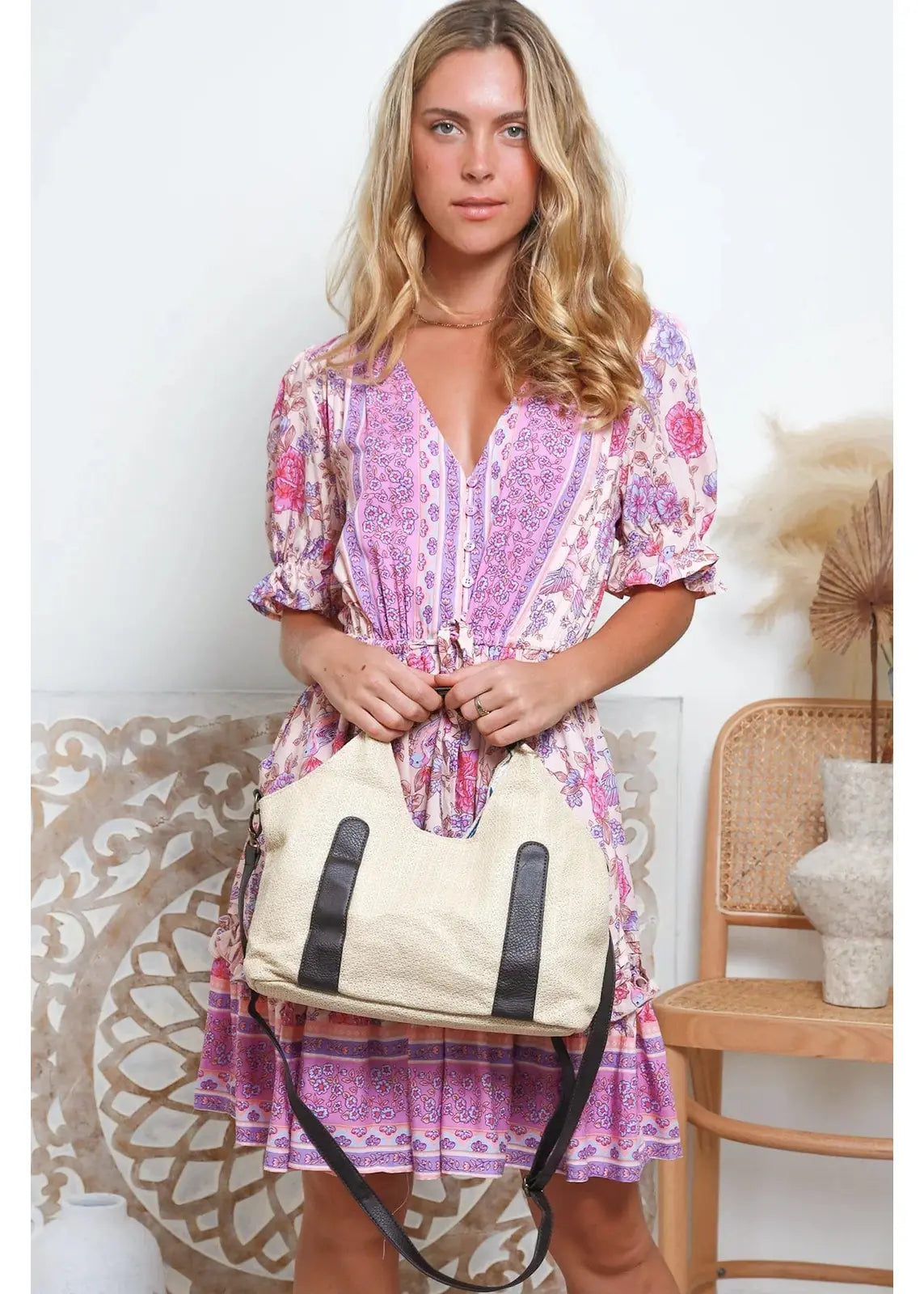 A woman wearing a pink and purple patterned dress holds a beige woven handbag with black accents and an adjustable black strap. The handbag has a casual, bohemian style.