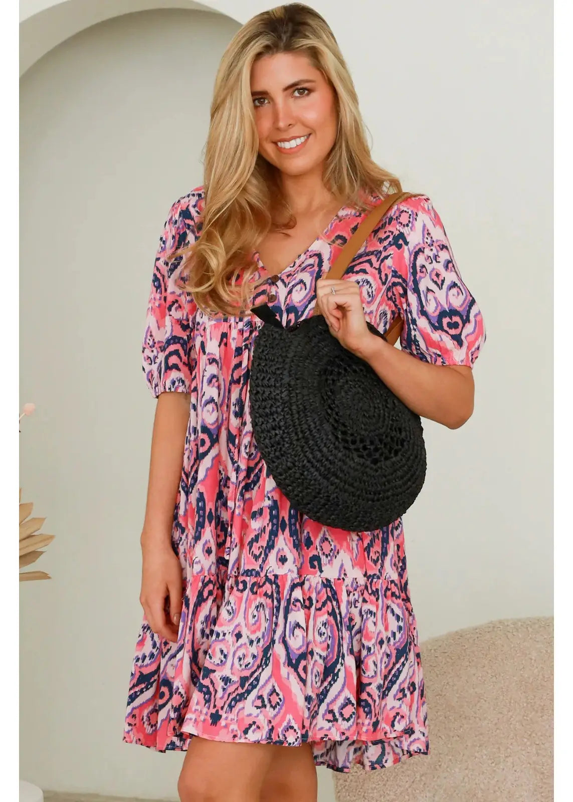 A woman wearing a pink and purple patterned dress holds a large, round black woven straw handbag with tan leather straps. The handbag has a circular, textured design, adding a stylish and bohemian touch to her outfit. She is smiling and holding the bag in front of her, highlighting its unique and fashionable look.