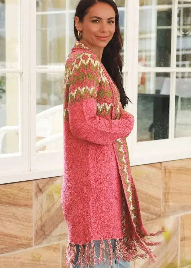 A woman wearing a long, open-front cardigan with a pink base and intricate boho-inspired patterns in shades of green and white. The cardigan features a fringed hem and is paired with a white top and blue jeans, creating a cozy and stylish look perfect for a casual day out.