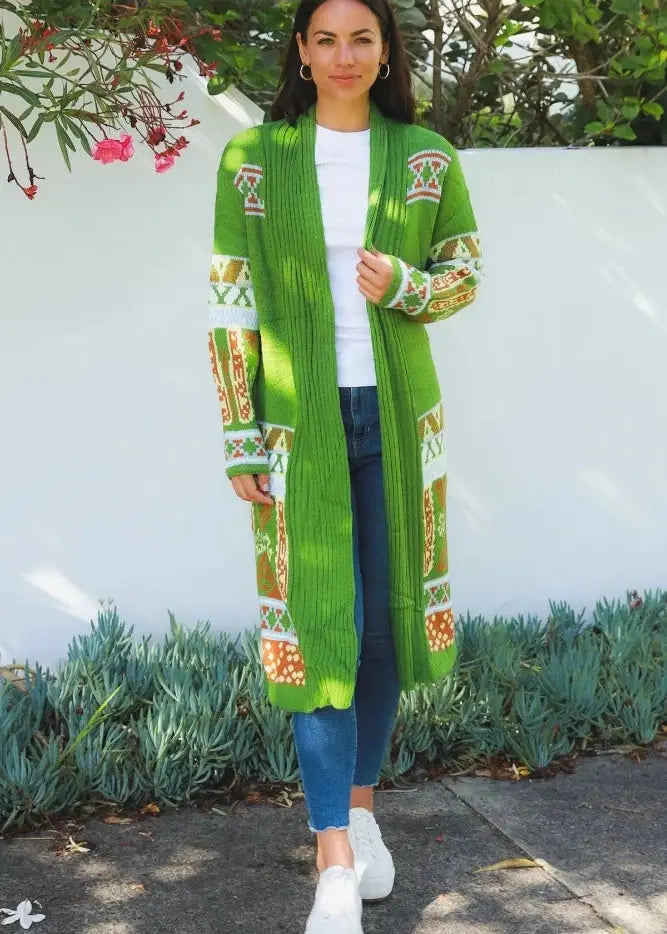 A woman stands outdoors wearing a striking below the knee knit cardigan with vibrant shades of green, detailed with white, orange, and yellow Aztec patterns along the trim and cuffs. Her look is casual-chic, paired with a white tee, blue jeans, and white sneakers. A blue cap and gold hoops complete her outfit. 