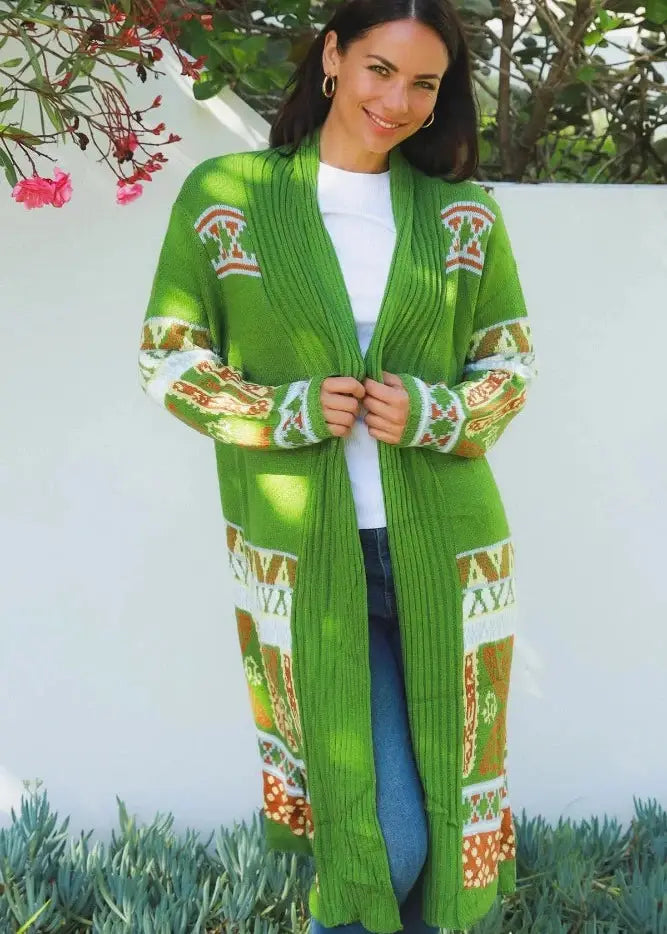 A woman stands outdoors wearing a striking long knit cardigan with vibrant shades of green, detailed with white, orange, and yellow Aztec patterns along the trim and cuffs. Her look is casual-chic, paired with a white tee, blue jeans, and white sneakers. A blue cap and gold hoops complete her outfit. 