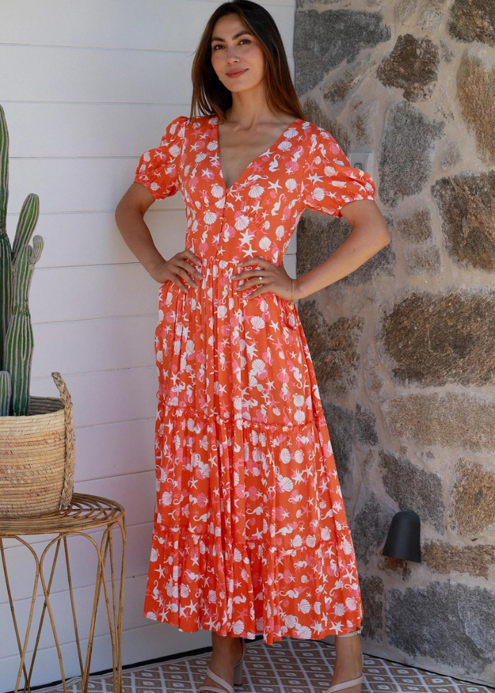 Woman wearing a orange/coral dress with a Vibrant and playful white seashell and starfish pattern, perfect for summer. The dress has a Deep V-neckline, which is A flattering cut that enhances your neckline. The Puff Sleeves Adds volume and a fun, trendy look. The Tiered Maxi Skirt Flows beautifully with every step, providing both comfort and style. The Lightweight Fabric is ideal for warm weather, keeping you cool and comfortable.