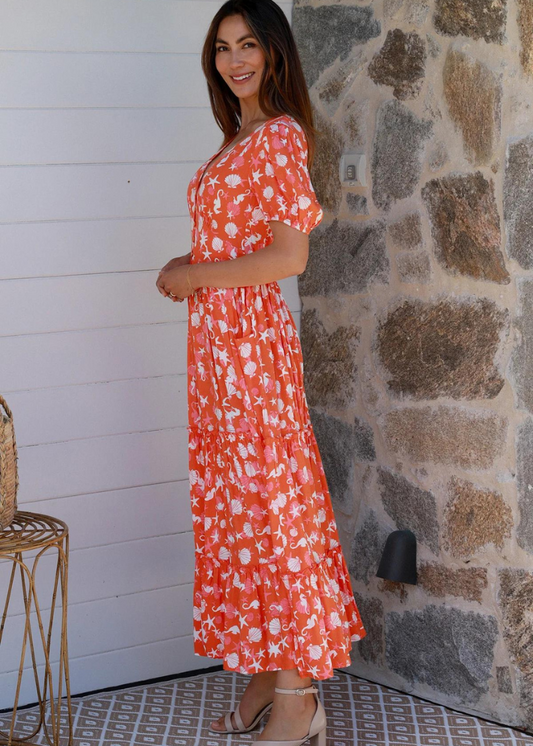 Woman wearing a orange/coral dress with a Vibrant and playful white seashell and starfish pattern, perfect for summer. The dress has a Deep V-neckline, which is A flattering cut that enhances your neckline. The Puff Sleeves Adds volume and a fun, trendy look. The Tiered Maxi Skirt Flows beautifully with every step, providing both comfort and style. The Lightweight Fabric is ideal for warm weather, keeping you cool and comfortable.