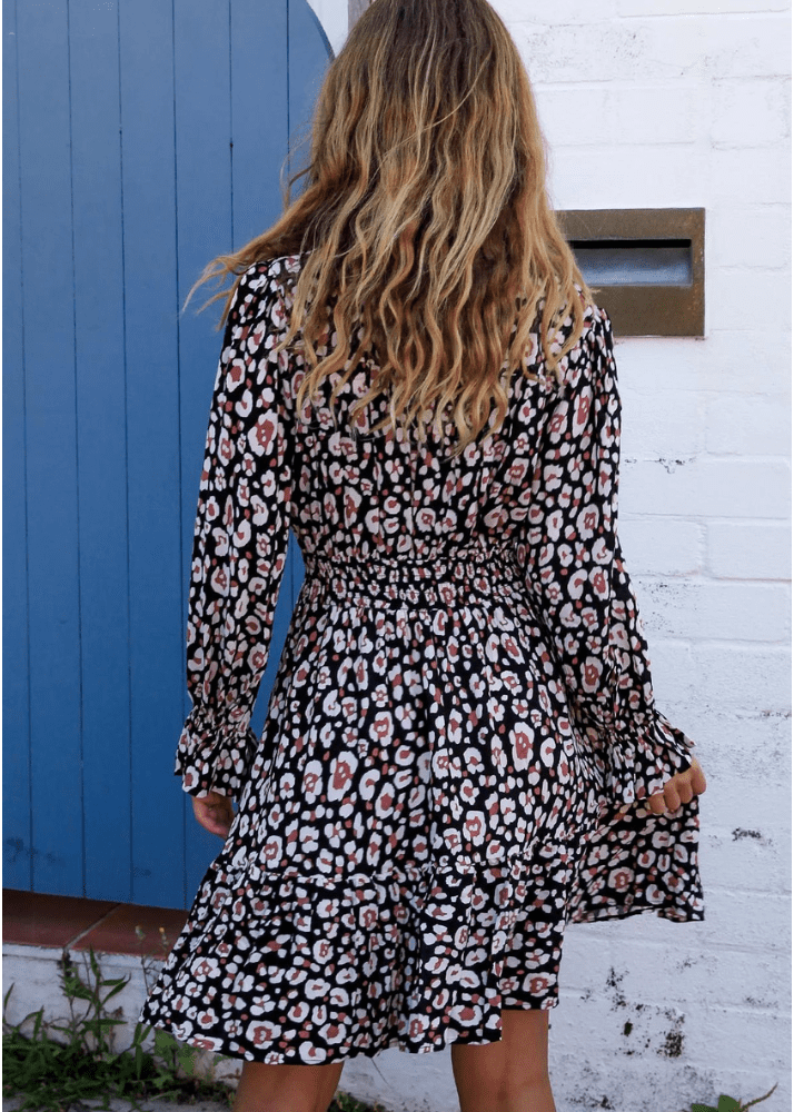 A woman wearing the Kelsey Mini dress. The dress is a long-sleeved black mini dress with a dusty pink and white leopard print pattern. The dress features a V-neckline and a cinched waist, creating a flattering silhouette, with a skirt that flares out gracefully at the hem.