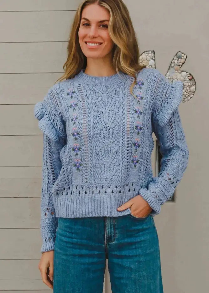 A woman wearing a light blue, knit sweater featuring intricate patterns and floral embroidery. The boho sweater has ruffled details on the shoulders and is paired with high-waisted blue jeans. She is smiling and standing against a neutral-colored wall, creating a casual and cozy look.