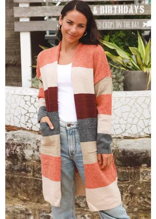 A woman wearing a cozy, multi-coloured striped knit cardigan in earth tones of cream, rust, and grey that features pockets styled effortlessly with a white t-shirt and light-wash denim jeans.